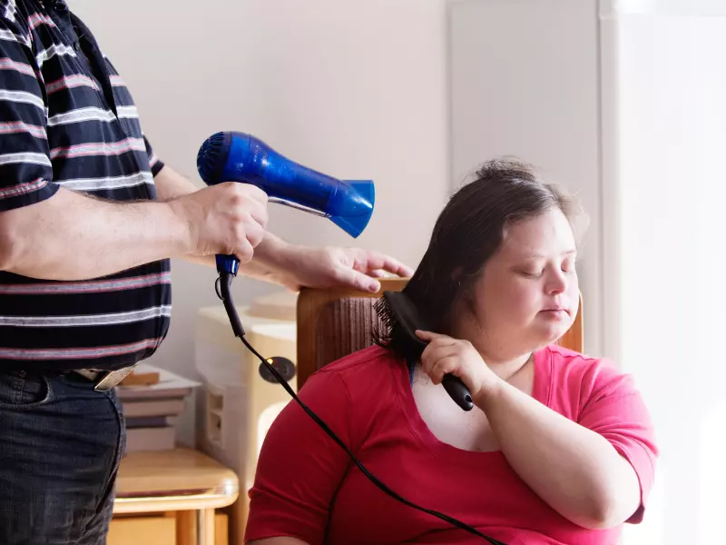 Disabled girl with down syndrome sitting and brushing hair while carer helps to blow dry